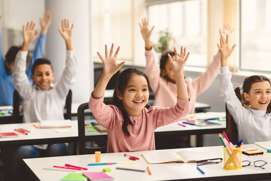 Elementary School and People Concept. Diverse excited group of emotional happy junior school kids sitting at desks in classroom and raising hands, having fun, studying with pleasure