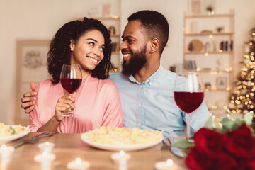 Happy Loving Family. Smiling african american couple sitting at table, holding glasses, drinking red wine, having gourmet meal at restaurant or at home. Young black guy embracing his lady in dress