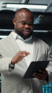 Vertical video Smiling admin using tablet in in data center maintaining infrastructure for workloads. Joyful IT professional in server farm utilizing high tech server clusters for parallel processing