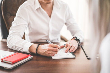 Man in white shirt writing in a notebook on a desk with laptop and red notebook present nearby