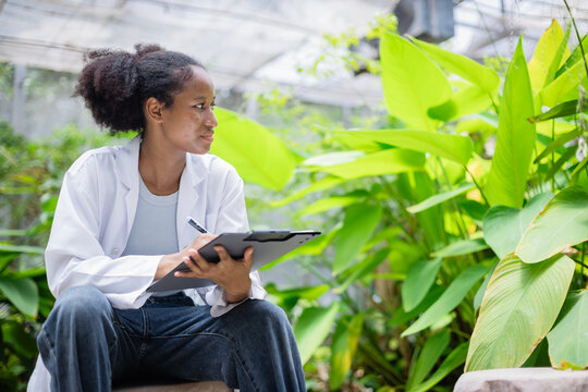 Young scientist in white coat is conducting agricultural research in farm environment while holding clipboard and observing plants with focus and care
