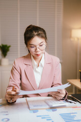 A focused young Asian businesswoman in a pink blazer works diligently at her desk, using a laptop...
