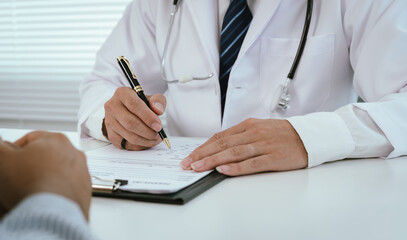 A doctor, wearing a white lab coat, holds a clipboard and pen, pointing towards it while interacting with a patient, indicating explanation or signature.