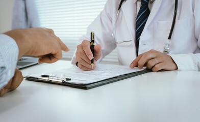 A doctor, wearing a white lab coat, holds a clipboard and pen, pointing towards it while interacting with a patient, indicating explanation or signature.