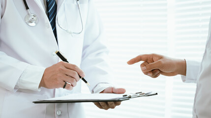 A doctor, wearing a white lab coat, holds a clipboard and pen, pointing towards it while...