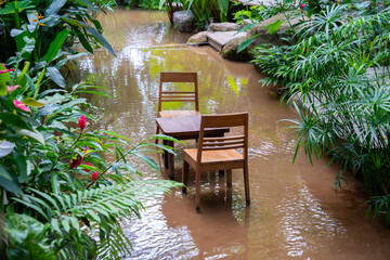 Wooden table and chairs are partially submerged in flooded garden, surrounded by lush greenery and vibrant flowers