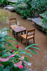 Wooden table and chairs submerged in flooded garden, surrounded by lush greenery and vibrant flowers