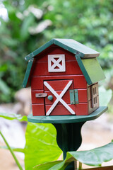 Small, red barn style mailbox with green roof stands in garden setting, surrounded by lush greenery