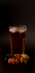 Tall glass of grape juice mixture with ice on black background, ripe bunches nearby.