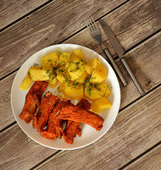 Marinated pork ribs baked in the oven and young boiled potatoes on a dark wooden table, cutlery nearby.