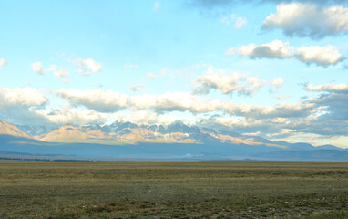Endless steppe in the shade and snowy mountain range in the clouds illuminated by the rising sun on a summer morning.