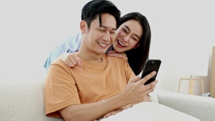 Asian Happy couple smiling looking at phone together relaxing on sofa at home cozy joyful romantic moment leisure lifestyle concept relationship bonding love happiness relaxation technology connection - Powered by Adobe