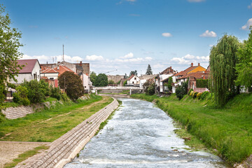 Kolubara River winds through the centre of Valjevo, Serbia, with low bridges and red-tiled houses lining grassy embankments under a bright spring sky, capturing small-town life along the water.