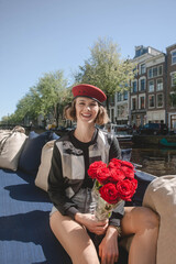 Beautiful cute woman wearing military red beret smiling into camera and holding red roses, while sitting on boat on canals in Amsterdam, Nederland