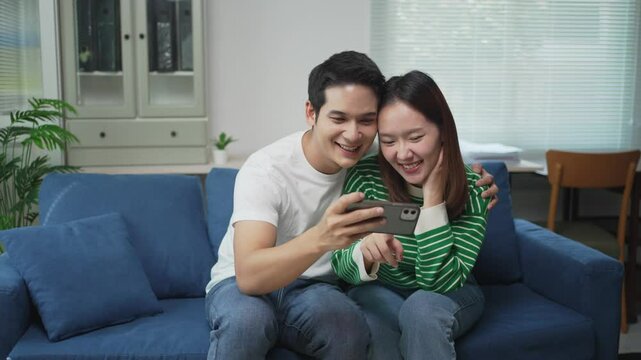 Young asian couple enjoying funny online videos while relaxing together on comfortable sofa, sharing lighthearted moments in modern living room