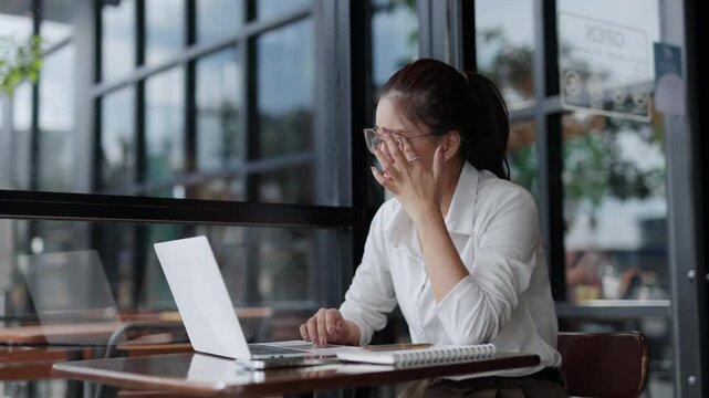 Office syndrome concept, young Asian businesswoman taking off glasses and rubbing tired eyes after long hours working on laptop computer in cafe