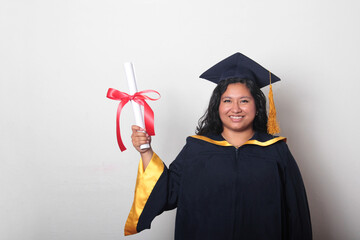 Young brunette Latina woman dressed in a cap and gown shows her diploma proudly, satisfied and...