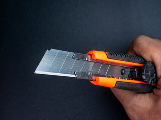 Close-up of hand holding metal utility knife with orange-black handle and sharp blade, ideal for office or work tools illustration.