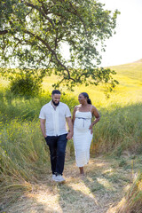 In a radiant, sunlit field filled with vibrant greenery, a joyful and expecting couple strolls hand in hand, cherishing their love and the exciting journey of welcoming their baby soon