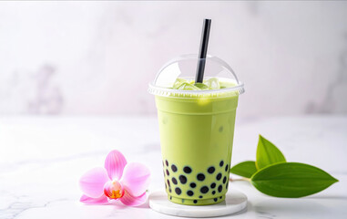 Creamy matcha bubble tea with black tapioca pearls in clear cup, accompanied by pink orchid and green leaves on marble surface