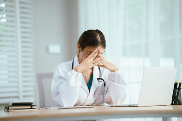 A concerned female doctor in a white lab coat and stethoscope holds her forehead, reading a document at her desk with a laptop.