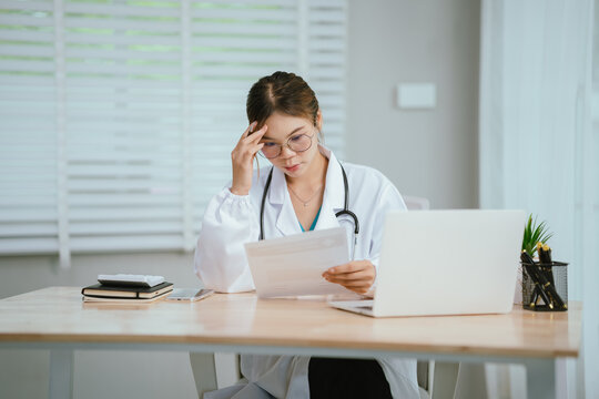 A concerned female doctor in a white lab coat and stethoscope holds her forehead, reading a document at her desk with a laptop.
