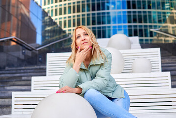 Fototapeta premium A woman is sitting on a bench in front of a building. She is wearing a green jacket and blue jeans