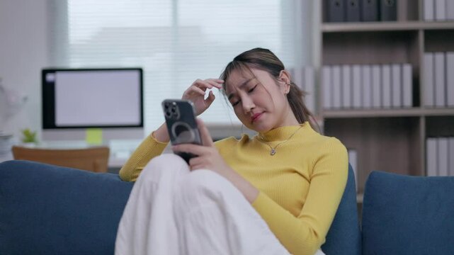 Stressed businesswoman experiencing emotional distress while reading upsetting message on smartphone, sitting dejectedly on comfortable sofa in home office environment