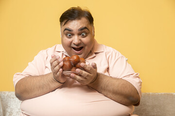 Fat indian man holding bowl of gulab jamun sweets. Overweight guy enjoy have craving for high...