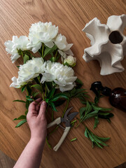 A woman collects a bouquet of white peonies on a wooden table, top view
