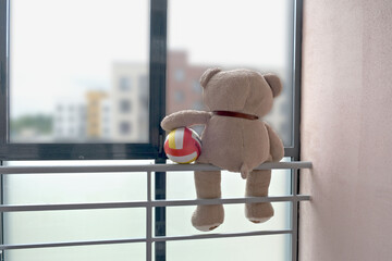 A teddy bear with a colorful ball sits perched on a balcony railing, looking out the window. The...
