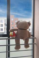 Teddy bear with a ball sits on the balcony railing against the blue sky outside the window