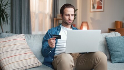 Cheerful man sitting on sofa and using laptop. Male sitting on couch and shopping online. Happy man...
