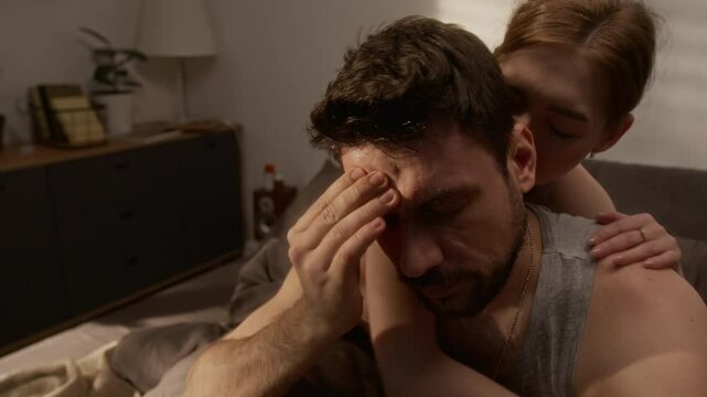 Close up shot of depressed adult man sitting on bed and rubbing face suffering from insomnia and panic attacks with caring wife embracing him