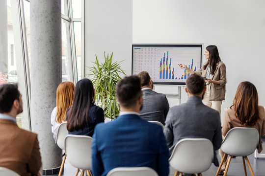 Young female analyst is standing near interactive board and showing charts and statistics to audience.