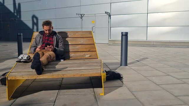 A man sits relaxed on a wooden bench at Arlanda Airport, casually smoking and scrolling on his smartphone&mdash;an unhurried moment in transit