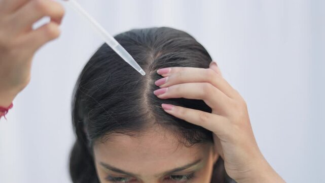 A young woman with a dropper applying hair serum on her hair root to improve hair growth in white background.