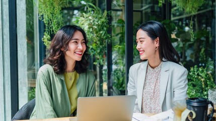 Happy conversation between senior and junior Asian businesswomen in green coworking space, casual professional attire, digital devices on desk