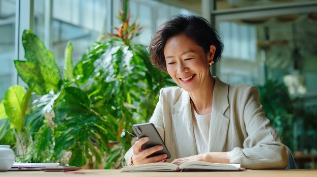 Middle-aged Asian businesswoman smiling warmly while checking smartphone, seated in sunny modern office with green plants, writing notes in open notebook