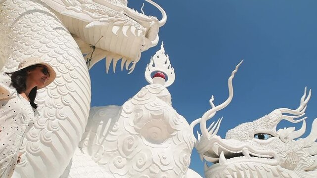 Woman tourist wear stylish white dress sightseeing explores iconic twin white dragons on temple stairs at Wat Huay Pla Kang in Chiang Raai, Thailand under clear blue sky. Famous travel phot spot