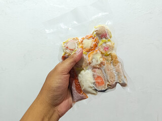 hand holding a clear vacuum-sealed package of frozen Siomay or Dim Sum, showcasing the various steamed dumplings with their fillings, against a plain white background, ready for cooking.