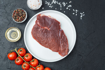 Raw beef steak on white plate with cherry tomatoes, pepper, salt and oil on black background