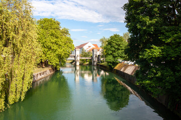 Obraz premium A serene and picturesque view of a tranquil canal or river in Ljubljana, on a bright, sunny day with a clear blue sky dotted with fluffy white clouds