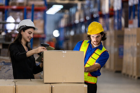 man in hard hat and safety vest assists women in scanning package while she records data during busy workday in a warehouse filled with shelves of goods.