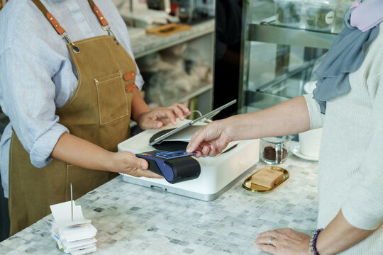 Asian senior female barista adult assisting customer using credit card for cashless payment at cafe counter demonstrating modern transaction in local family coffee shop, Concept small business