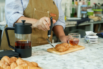 Asian senior female barista adult slicing croissant on wooden tray next to glass teapot and cup preparing order at cafe counter reflecting authentic service in small family coffee shop, small business