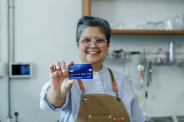 Cheerful Asian senior barista smiling, wearing apron, holding blue credit card in cafe, expressing readiness for digital payment, professionalism, joy, pride in modern small business.