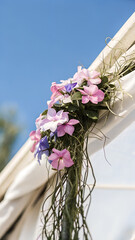 A floral arrangement with pink and purple flowers, green leaves and thin branches, hanging from a white fabric, with a blue sky in the background