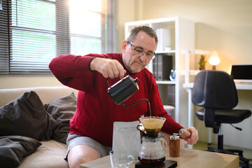 Senior man in red sweater making pour over coffee at home with a focused expression
