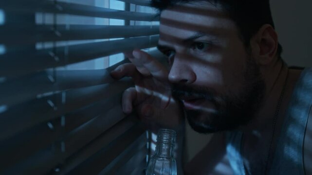 Close up shot of anxious adult man looking through blinds at night and drinking alcohol suffering from paranoia and mental health issues